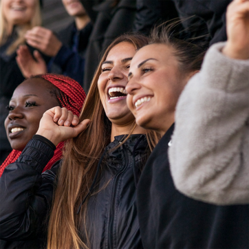 Three young women are joyfully celebrating, with wide smiles and laughter. The woman on the left has dark skin and red braids, while the others have light brown hair. They are wearing casual jackets and appear to be in a lively crowd, expressing excitement and happiness.