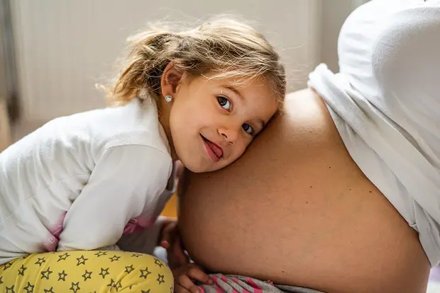A young girl hugs her pregnant mother's belly, smiling and playfully sticking out her tongue. The image captures a moment of affection and anticipation between the child and the mother, highlighting the bond before the arrival of a new sibling.