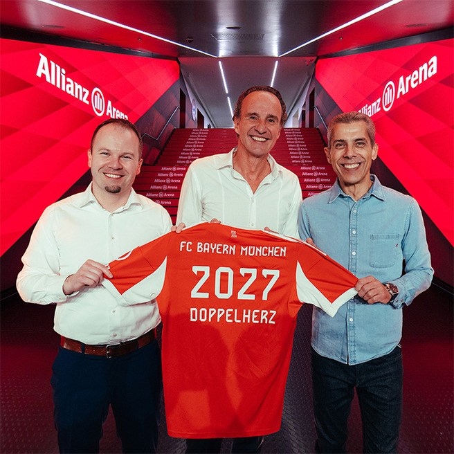 Three men stand together in a hallway at Allianz Arena, holding up a red FC Bayern München soccer jersey. The jersey features "2027" and "DOPPELHERZ" printed on the back. The background has a bright red design, emphasizing the team's branding.