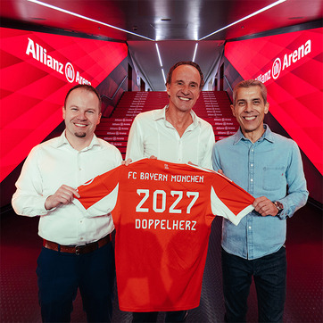 Three men stand together in a hallway at Allianz Arena, holding up a red FC Bayern München soccer jersey. The jersey features "2027" and "DOPPELHERZ" printed on the back. The background has a bright red design, emphasizing the team's branding.