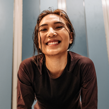 A young woman with wet hair smiles brightly at the camera, exuding joy and confidence. She is wearing a dark athletic top, and the background features soft blue tones, creating a vibrant and uplifting atmosphere.